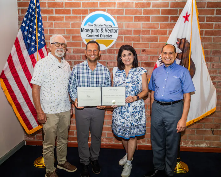 A group poses with a certificate at the San Gabriel Valley Mosquito and Vector Control District, U.S. flags in the background.
