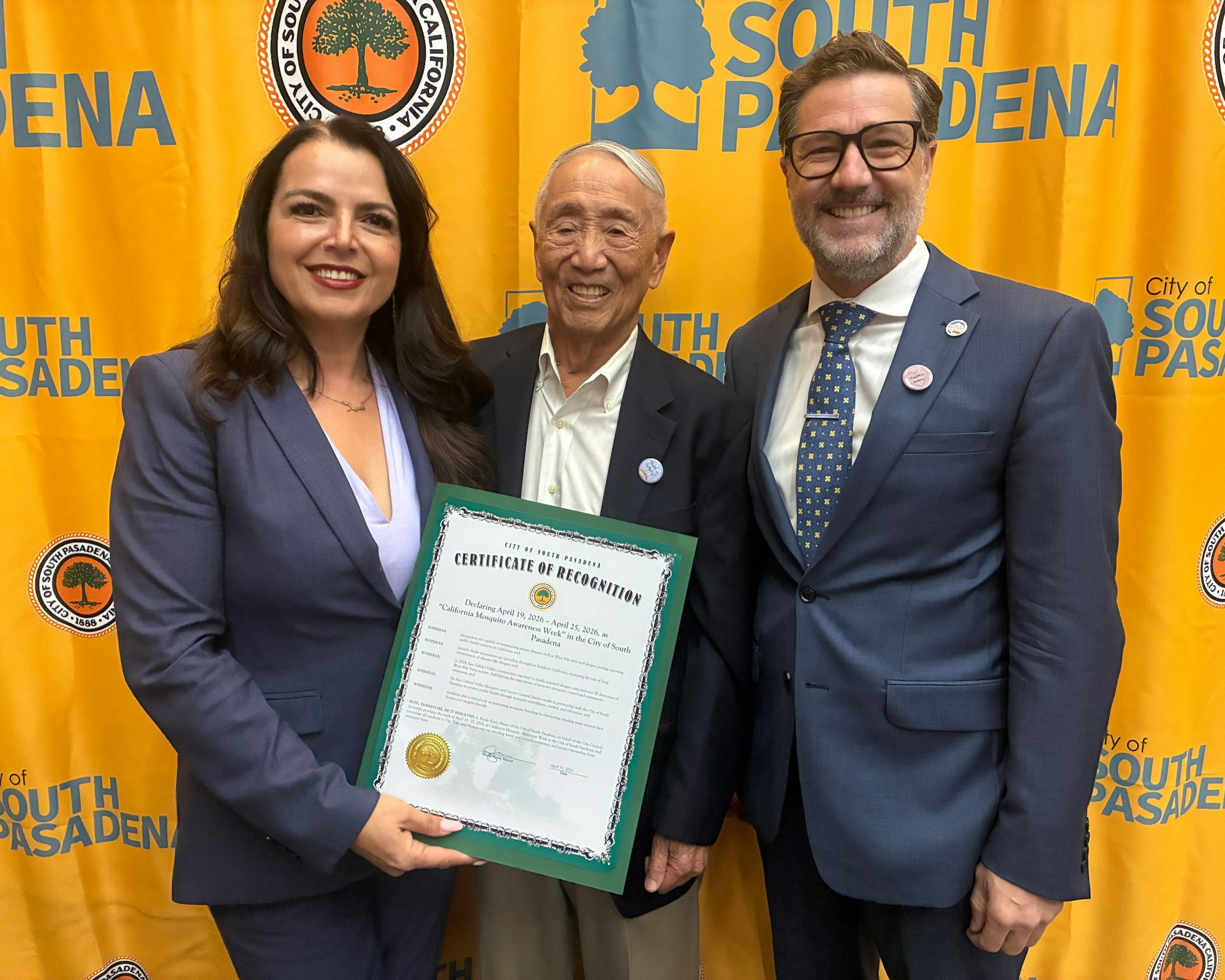 A recognition ceremony in South Pasadena featuring three people, holding a certificate of recognition against an orange backdrop.