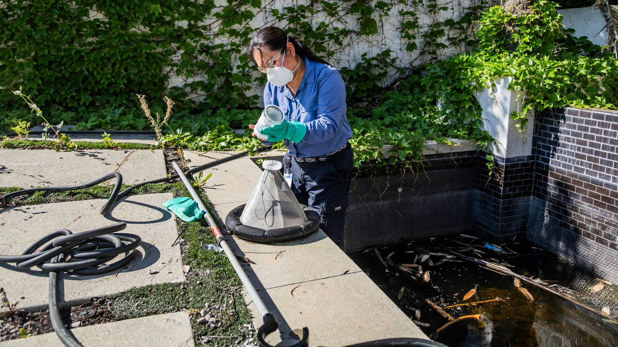 A person in protective gear is inspecting or collecting a sample near a stagnant water area surrounded by greenery.