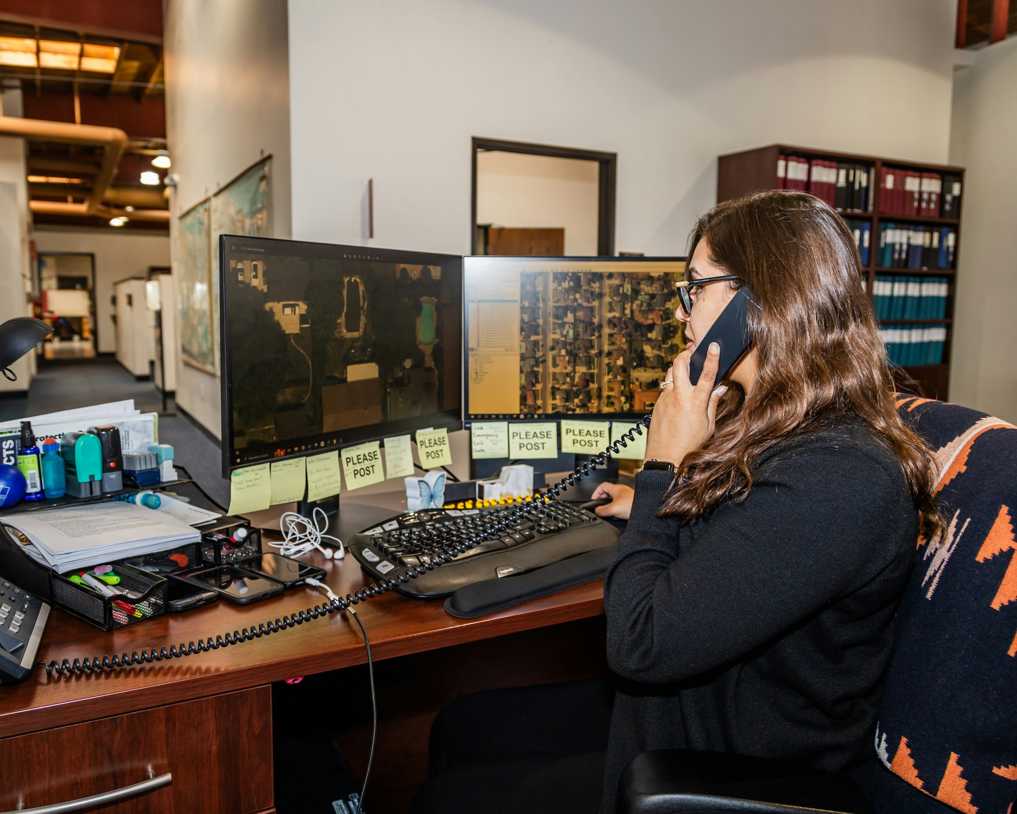 Woman on phone, working at a desk with dual monitors, sticky notes, and office supplies. Bookshelf in background.