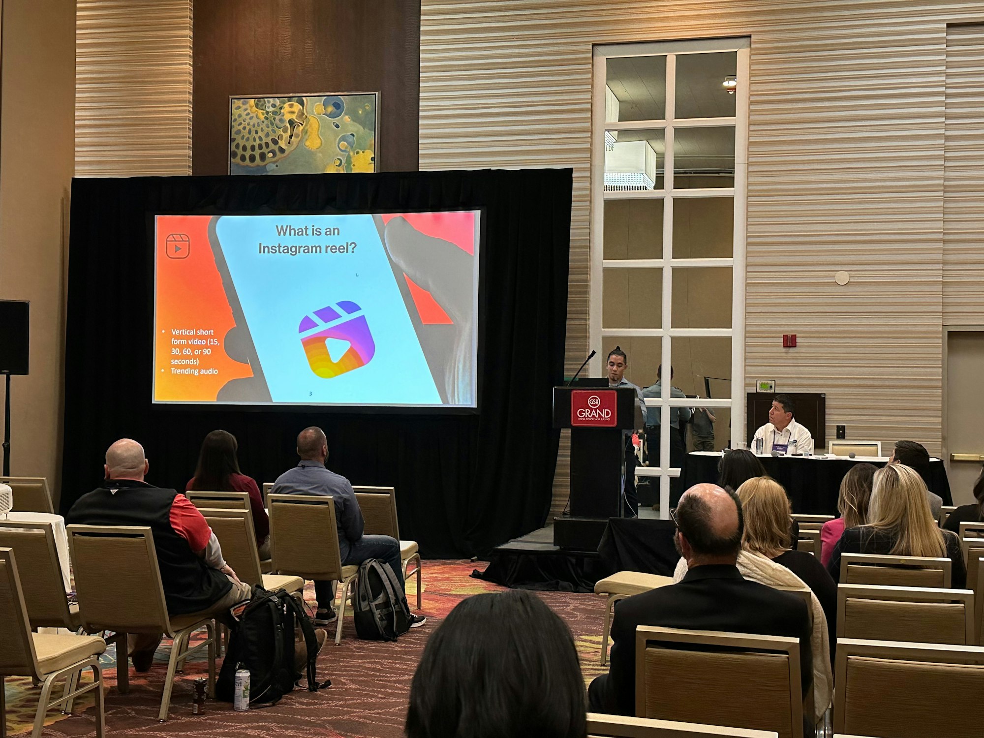 May contain: people, person, crowd, adult, female, woman, male, man, backpack, bag, architecture, building, classroom, indoors, room, school, audience, chair, furniture, electronics, speaker, electrical device, microphone, clothing, footwear, shoe, accessories, glasses, and lecture