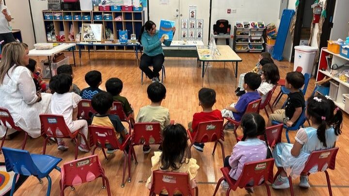 A teacher reads to children seated on small chairs in a classroom.