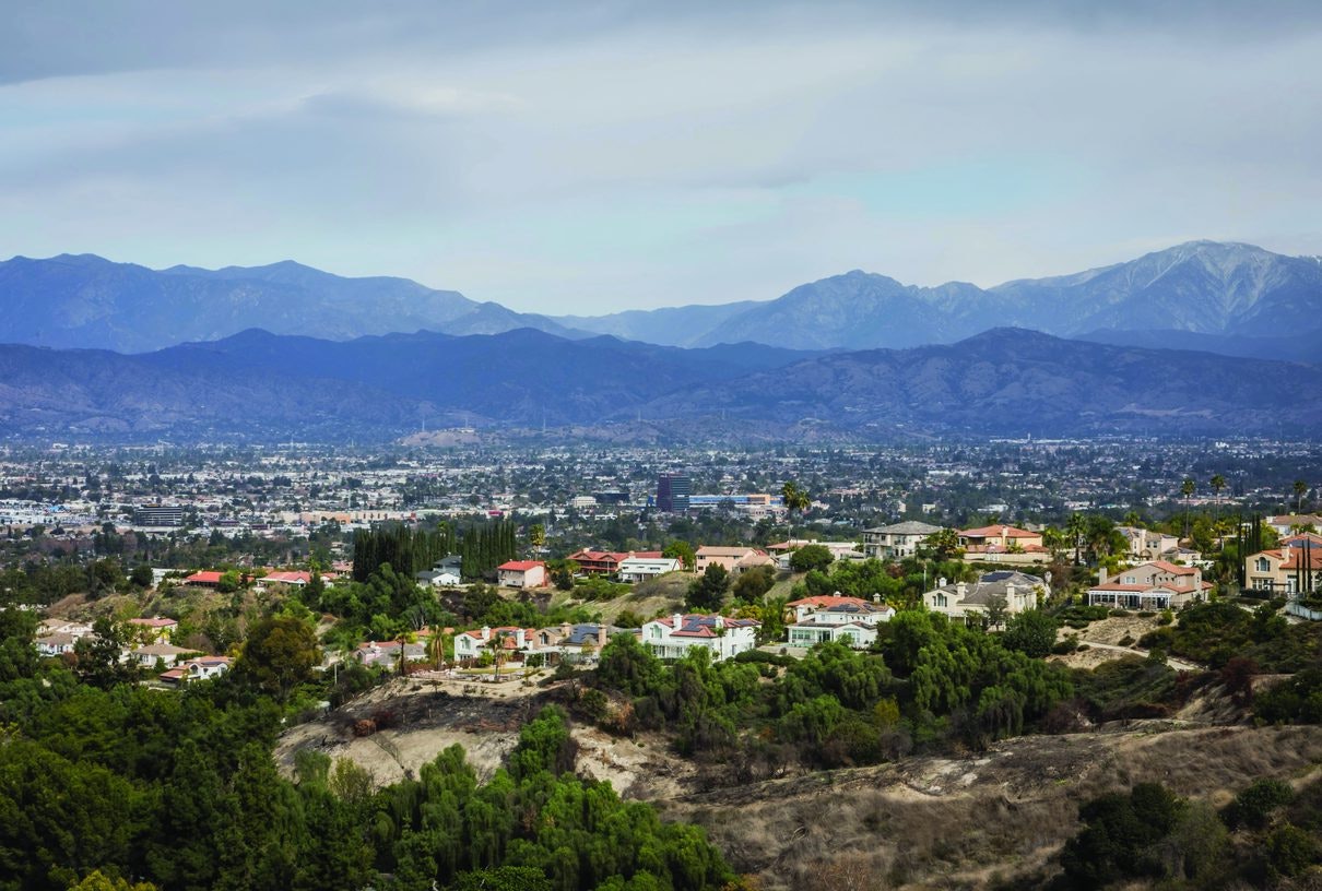 A scenic view of a suburban area with houses nestled among greenery, set against a backdrop of mountains and a cloudy sky.