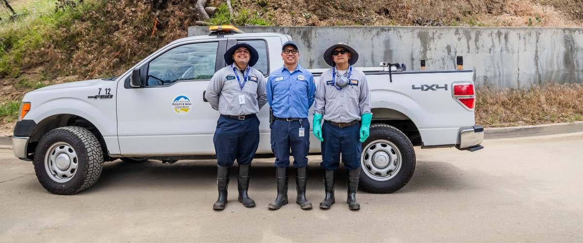 Three workers in uniforms stand together by a pickup truck, wearing hats and rubber boots, with a hillside in the background.