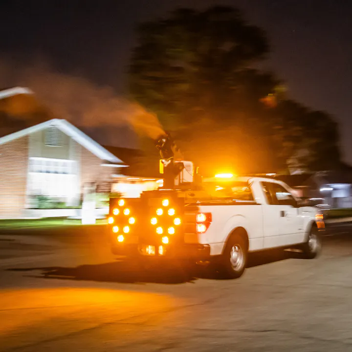 A white truck with bright lights is spraying something while driving at night near a residential area.