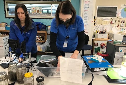 Two women in blue shirts are working at a table with documents, jars, and equipment in a classroom setting.