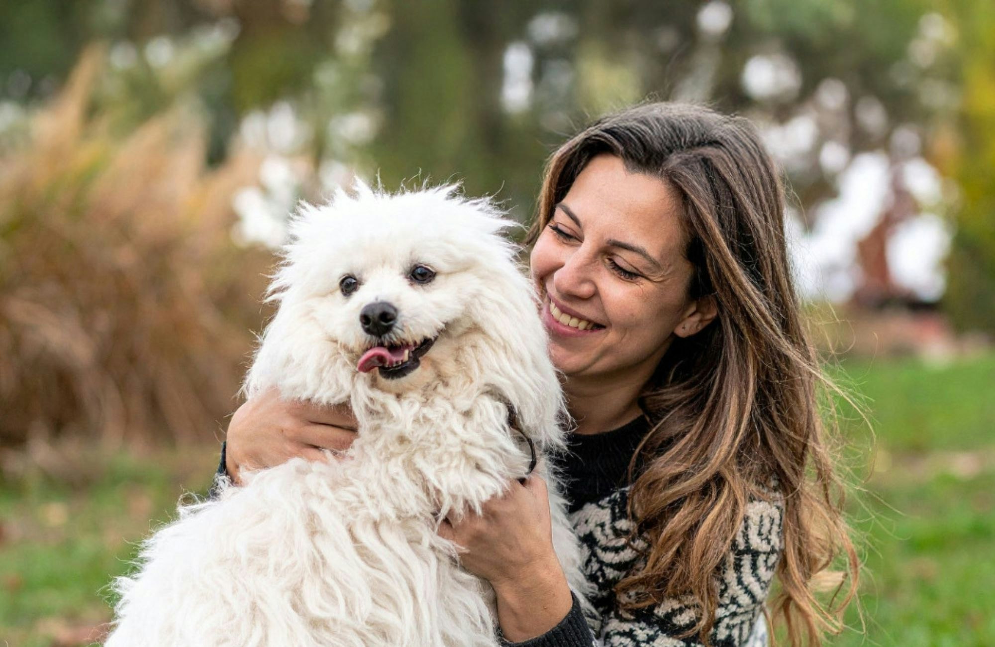 A woman happily embraces a fluffy white dog in a natural outdoor setting, showcasing a joyful bond between them.