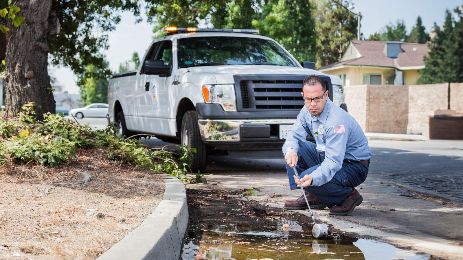 A technician in work attire inspects a puddle with a tool near a parked white truck on a suburban street.