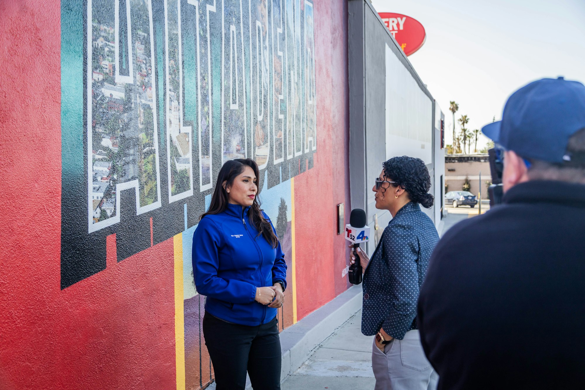 A woman in a blue jacket is being interviewed in front of a colorful mural, while another interviewer holds a microphone.