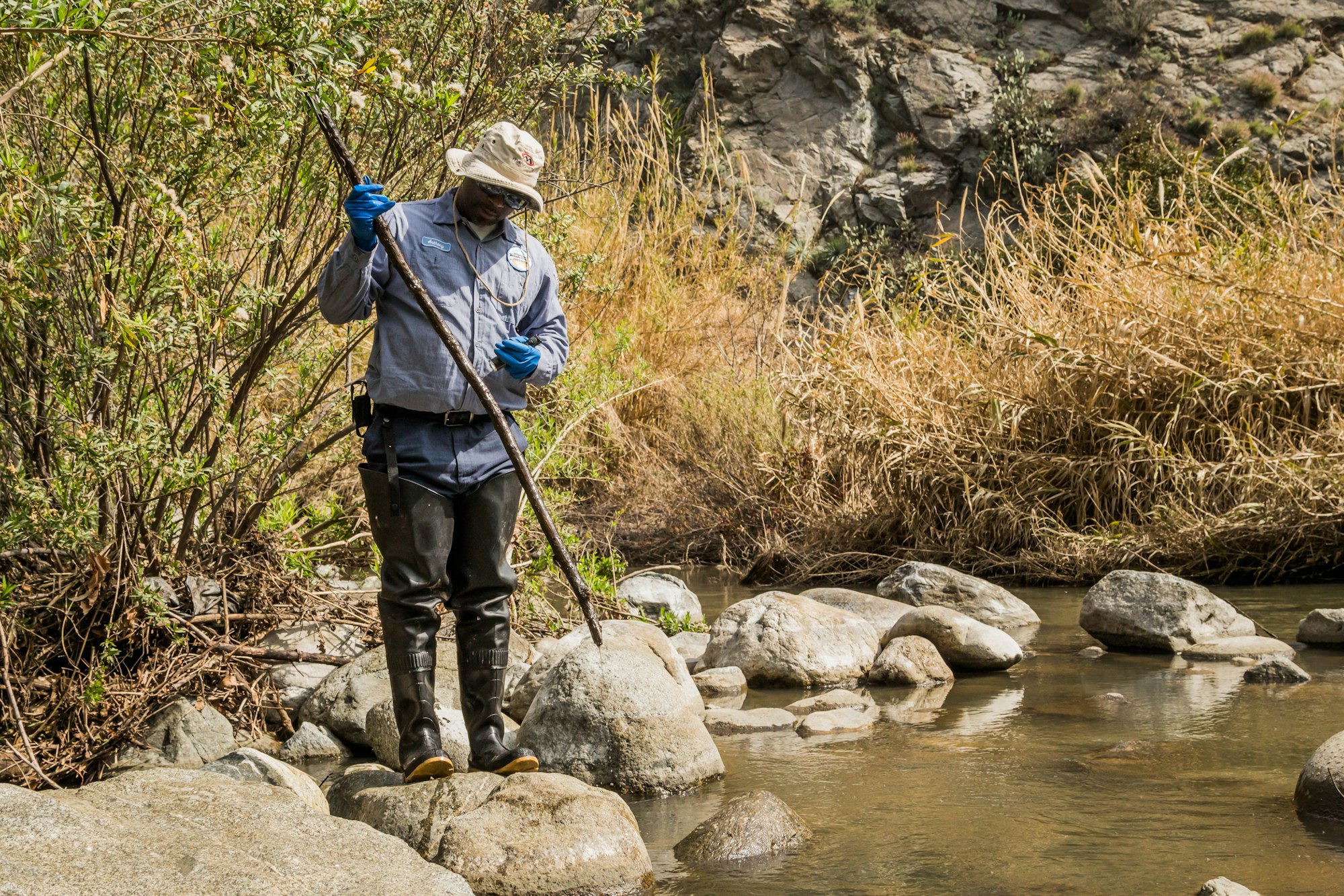 A person wearing boots and gloves stands by a stream, examining something with a stick amidst dry vegetation and rocks.