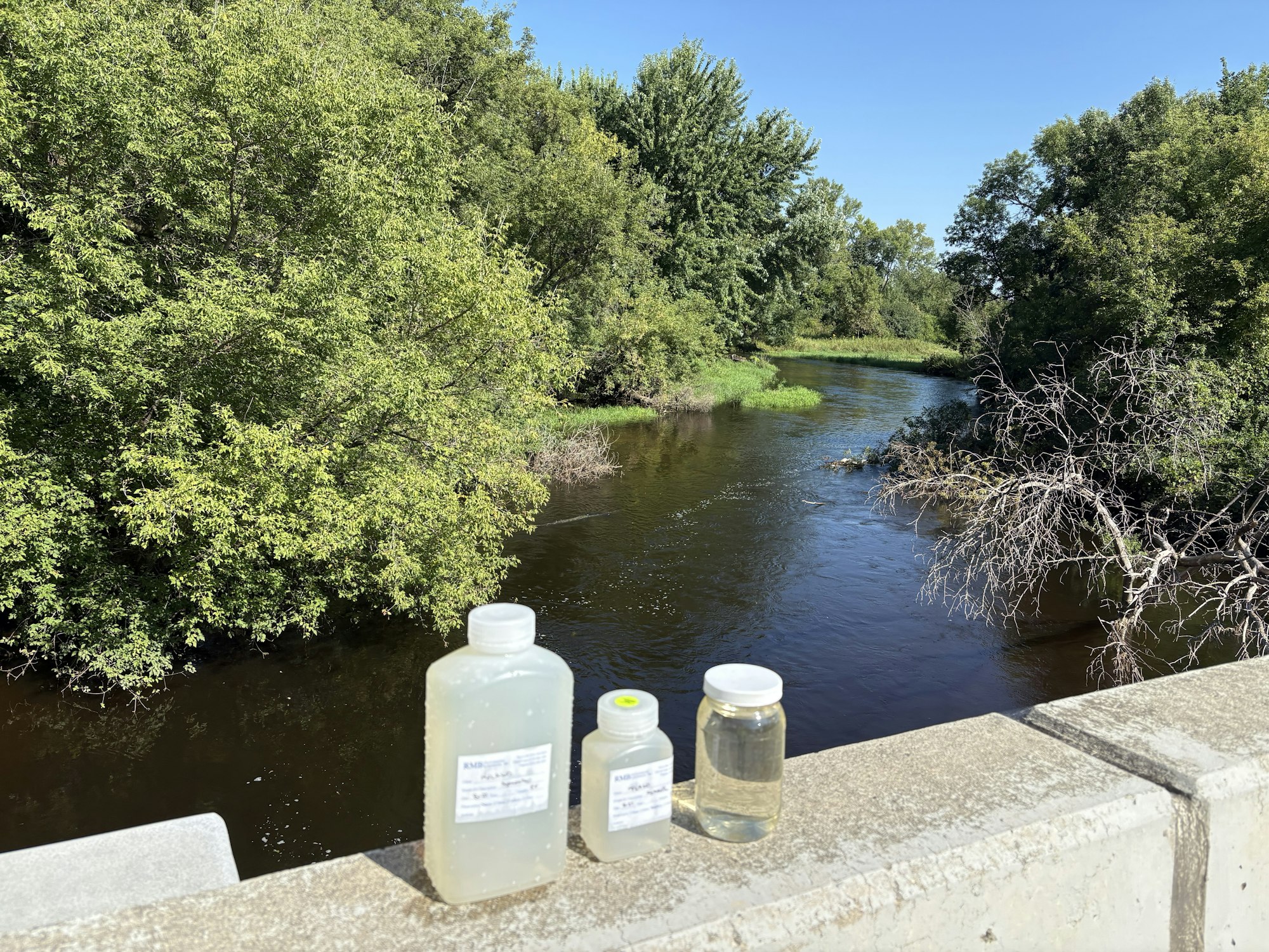 Bottles with labels on a ledge above a river with surrounding trees and clear blue sky.