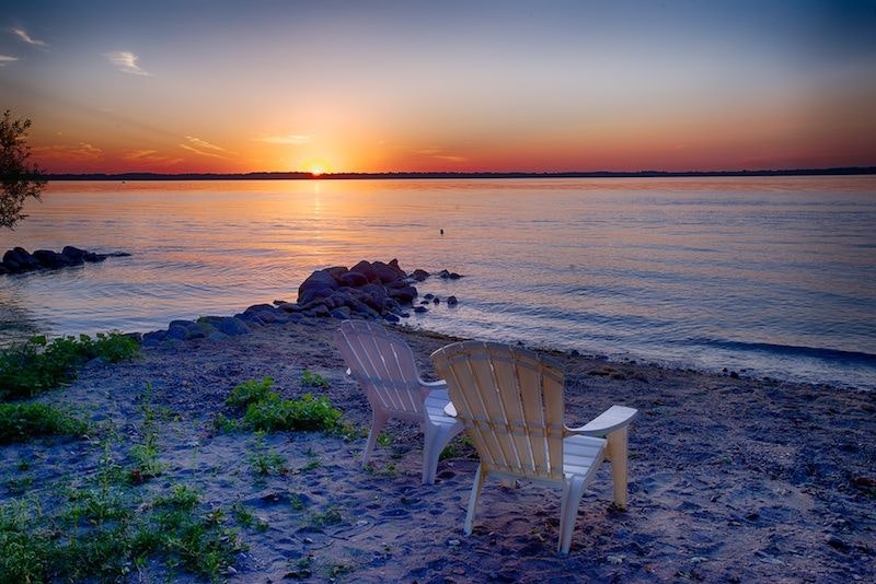 Sunset over a calm lake with two empty chairs on a sandy shore.