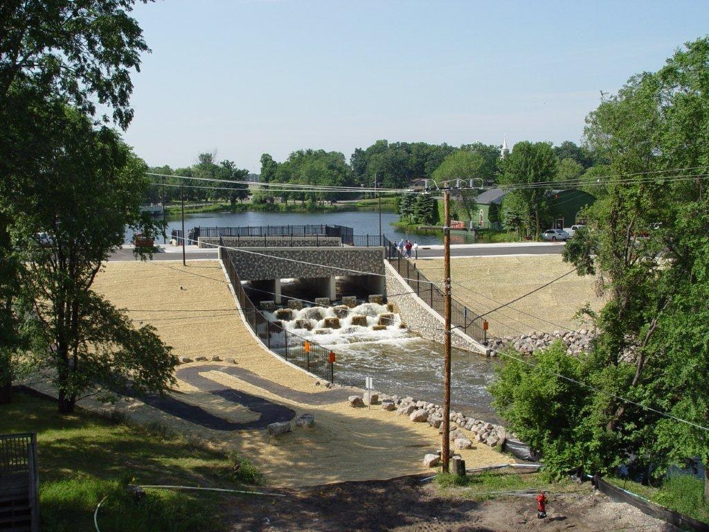 A small dam or weir with cascading water, surrounded by greenery and a distant lake.