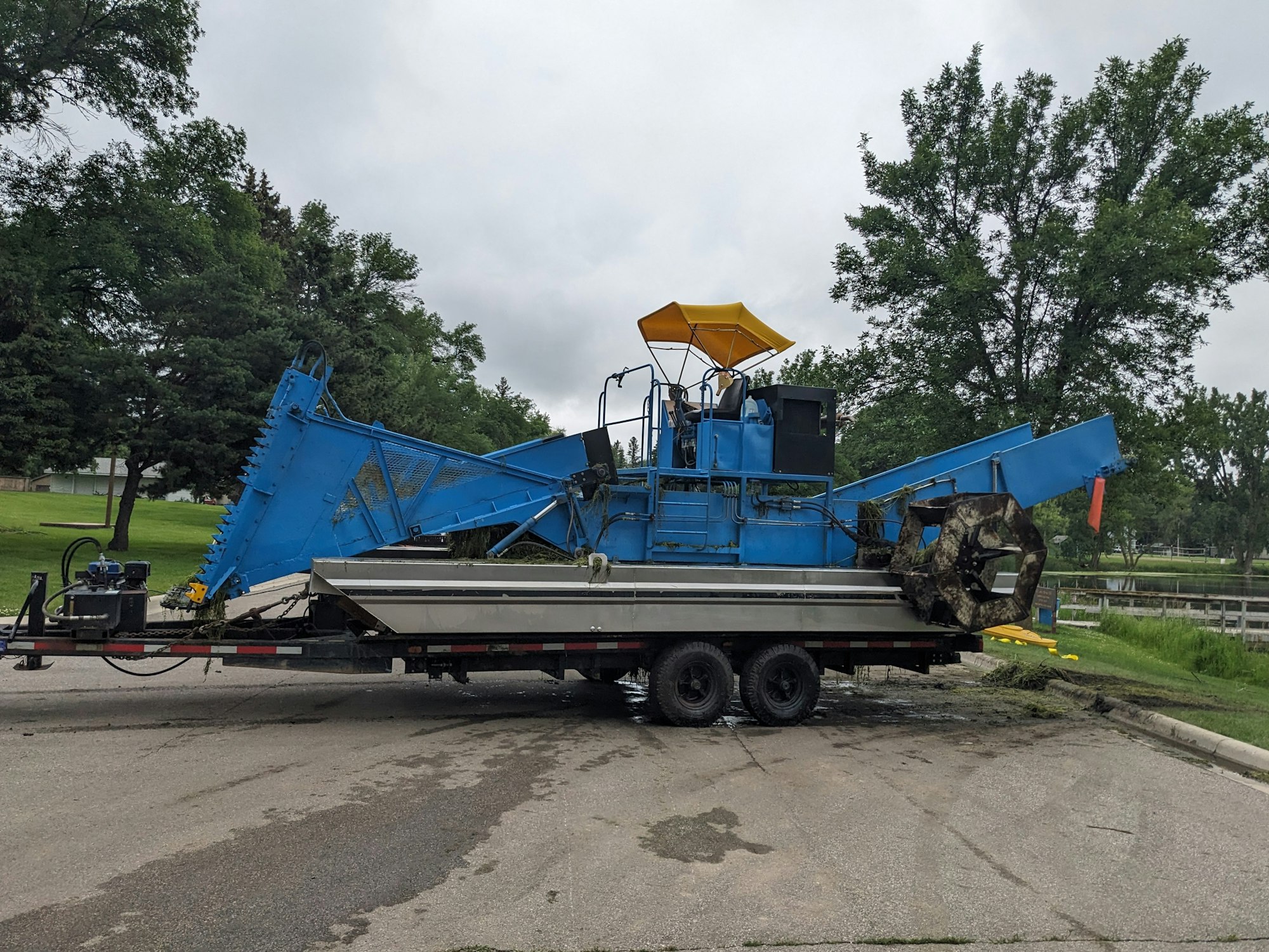 A blue industrial weed harvester mounted on a trailer, parked on an asphalt road.