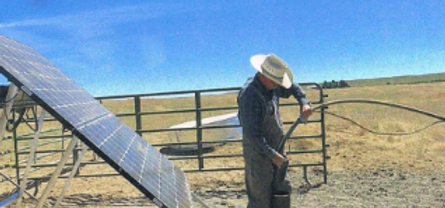 A person wearing a hat works near a solar panel and a fence in a rural setting.