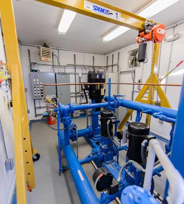 Industrial room with blue machinery, pipes, a hoist system, control panels, and electrical conduit on walls.
