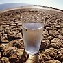 A glass of water sits on cracked, dry earth under a clear sky.