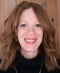 A person with long red hair smiling, wearing a dark turtleneck, against a wooden background.