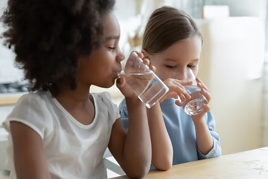 Two children are sitting at a table, drinking glasses of water.