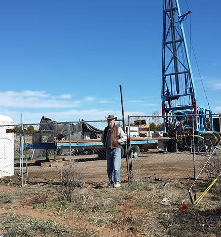 A person in a hat stands at a fence near an industrial site with a tall structure, possibly an oil rig, against a clear blue sky.