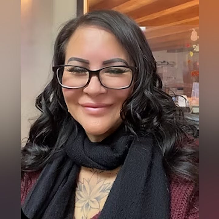 A person with glasses, dark hair, and a tattoo, wearing a black scarf and burgundy sweater, smiles at the camera.