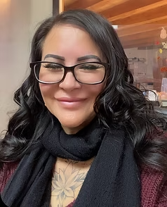 A person with glasses, dark hair, and a tattoo, wearing a black scarf and burgundy sweater, smiles at the camera.