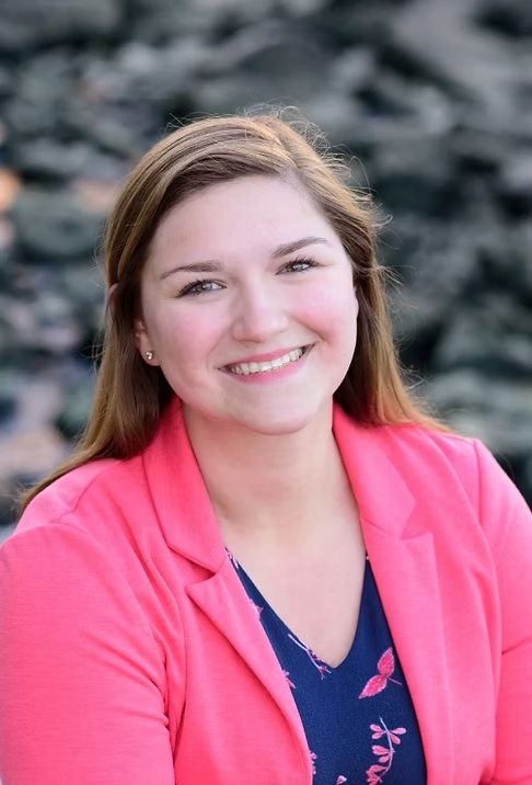 A person in a pink jacket smiling, with an outdoor background of rocks.