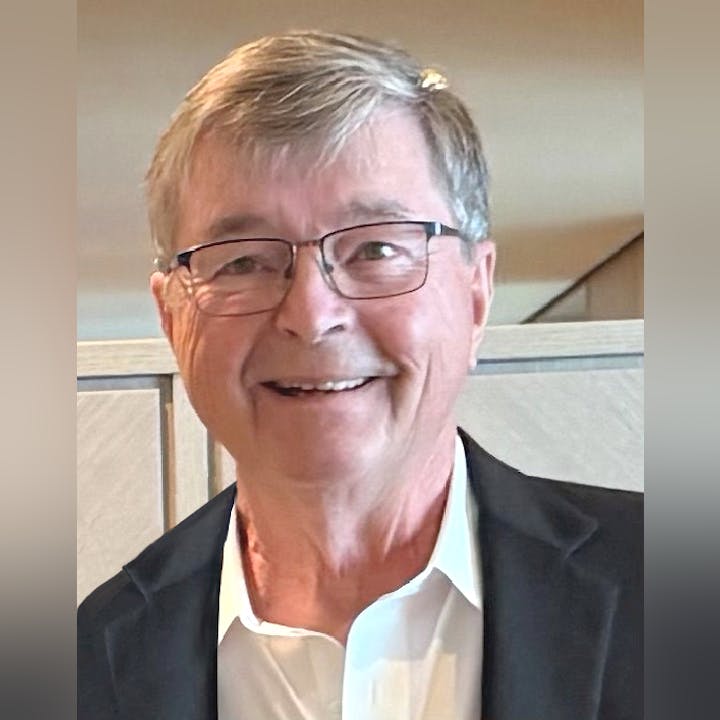 The image shows an older man with glasses, smiling, wearing a white shirt and a dark jacket, against a light background.
