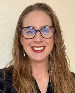 A person with long hair, glasses, and a black top smiles at the camera against a light background.