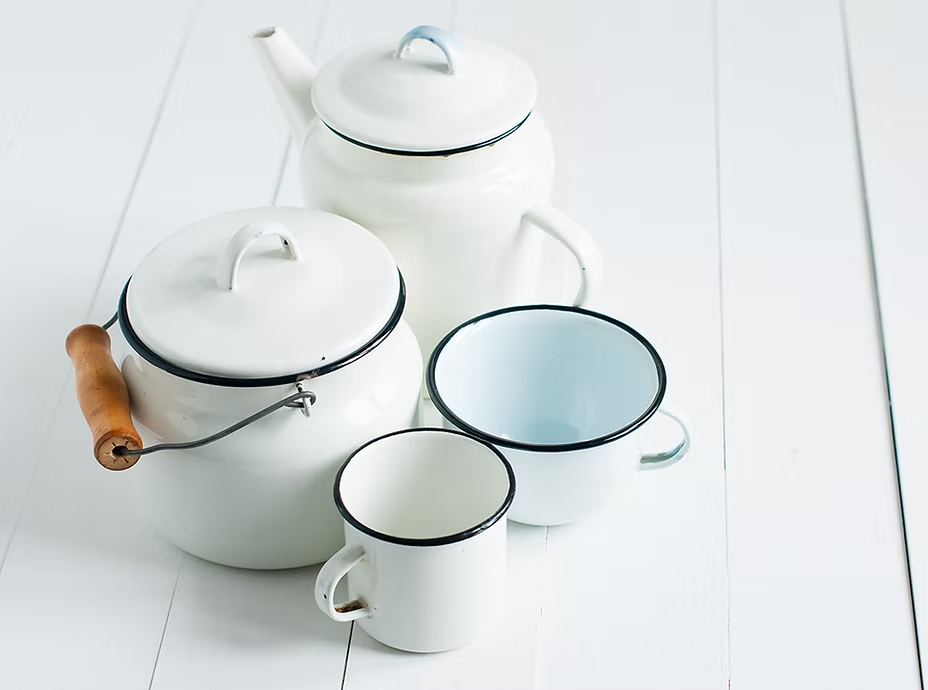 White enamel teapot, kettle, and two mugs with black rims on a white wooden surface.