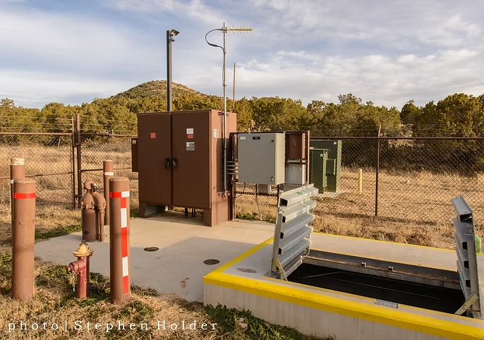 Fenced area with utility equipment, antenna, and an open hatch amidst a grassy landscape with trees and hills in the background.