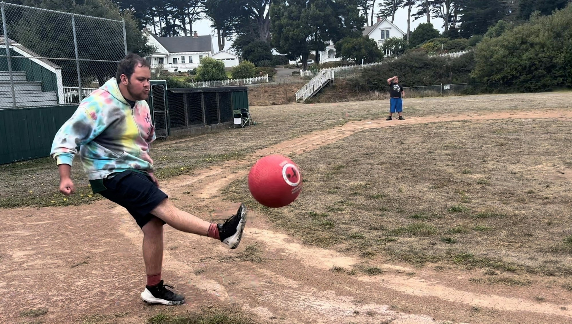 Man kicks a red ball in a park with another person watching in the background.
