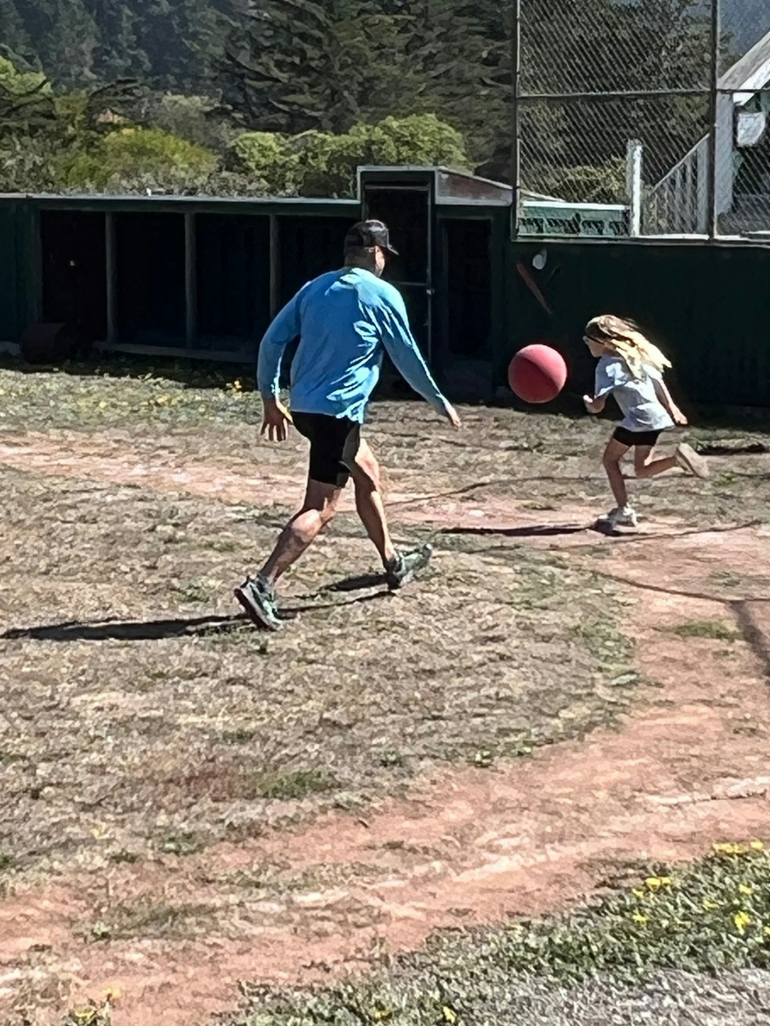 A man and a girl playing with a red ball outside near a fence and greenery.