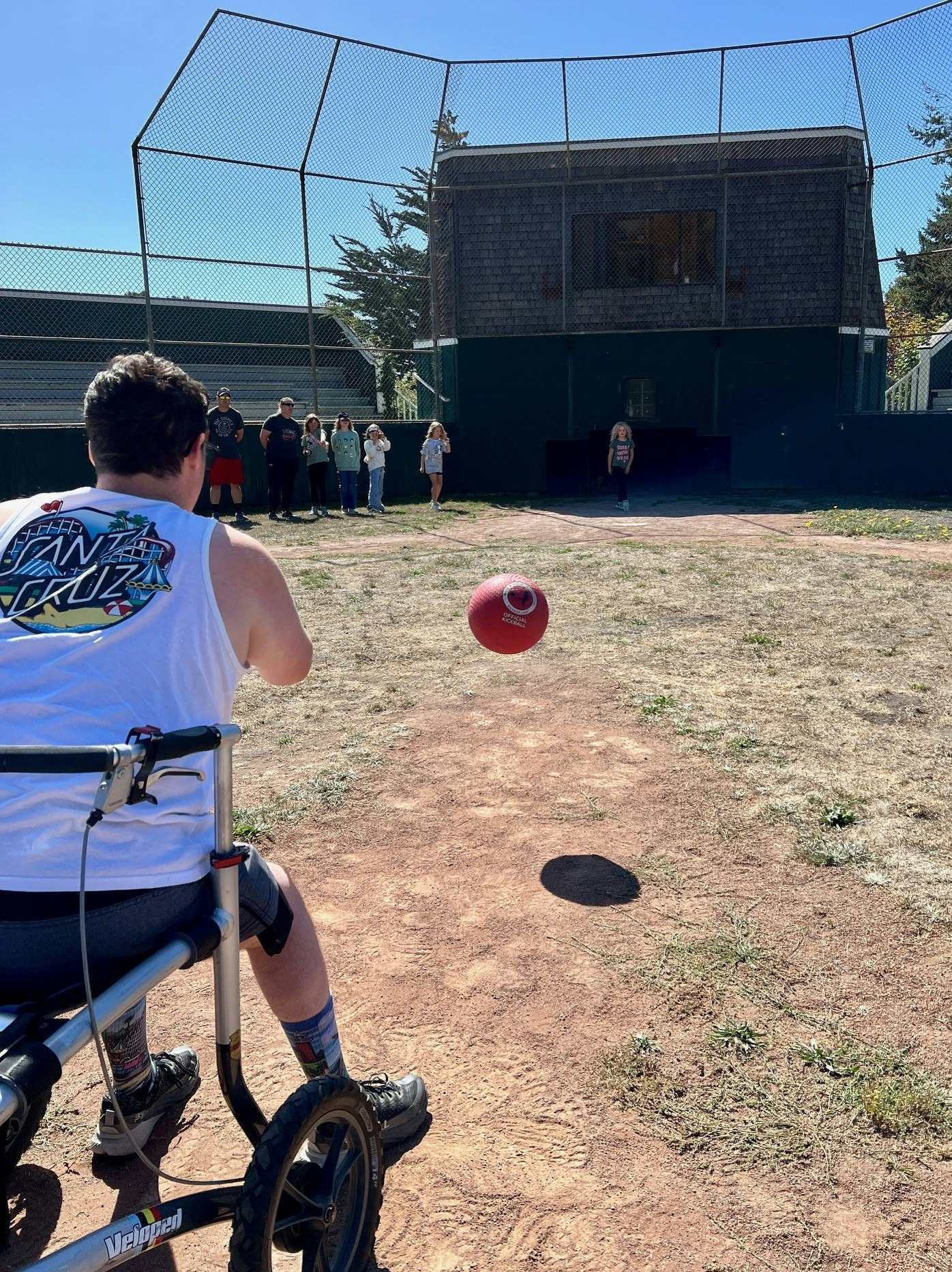 Person in a wheelchair playing catch with a red ball, onlookers in the background, sunny day at a ballpark.