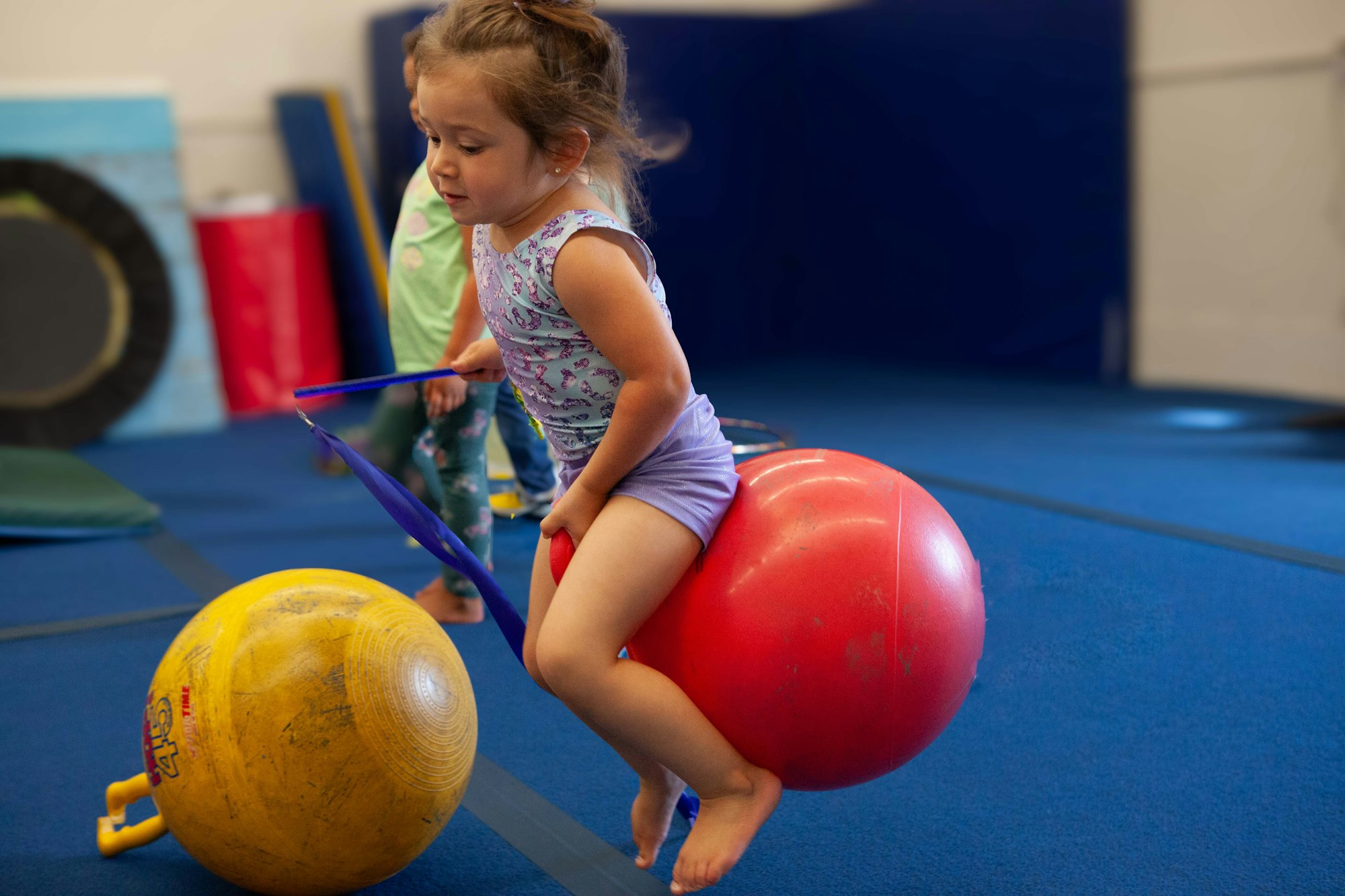 A child is sitting on a red gym ball with a hoop in hand, in a playful gym setting.