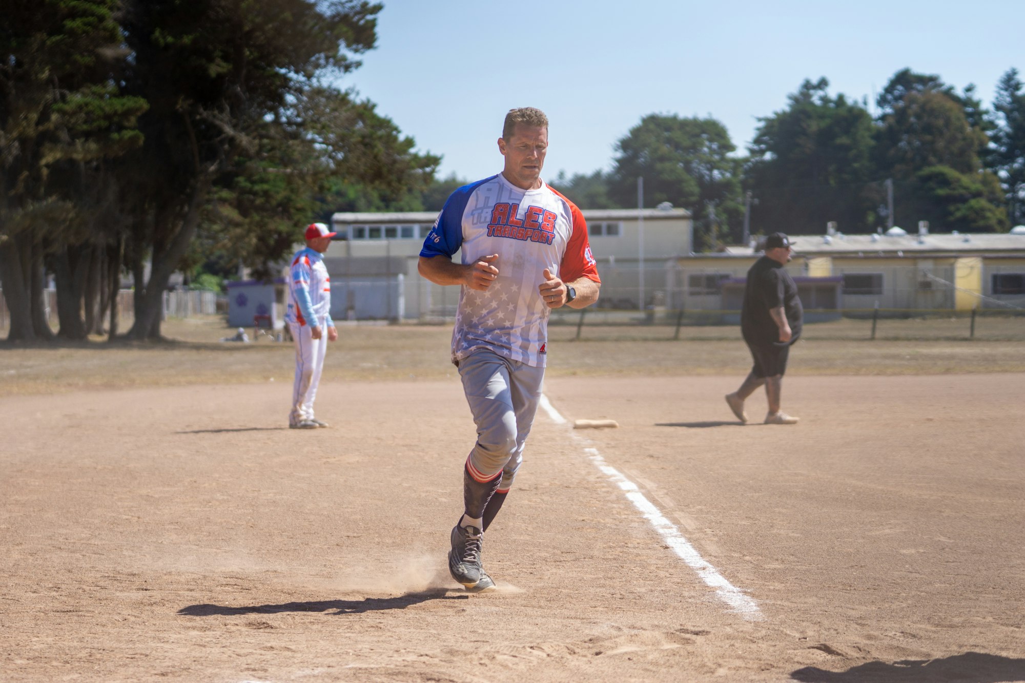 A man in a baseball uniform is running on a dirt field, with others and trees in the background.