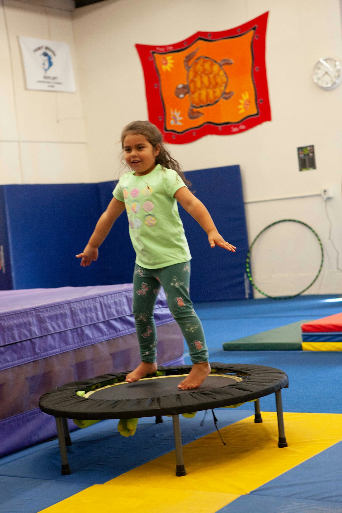 A child is bouncing on a mini trampoline in a gymnasium with colorful mats and a turtle banner on the wall.