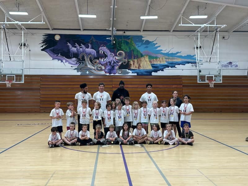 A group of children and adults in a gym with a dragon mural, all wearing similar shirts and some with medals.