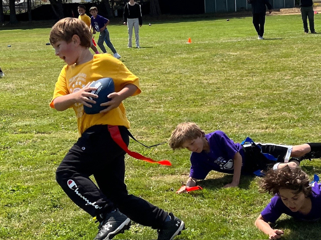A child in a yellow shirt runs with a football, evading two others in a game of flag football.