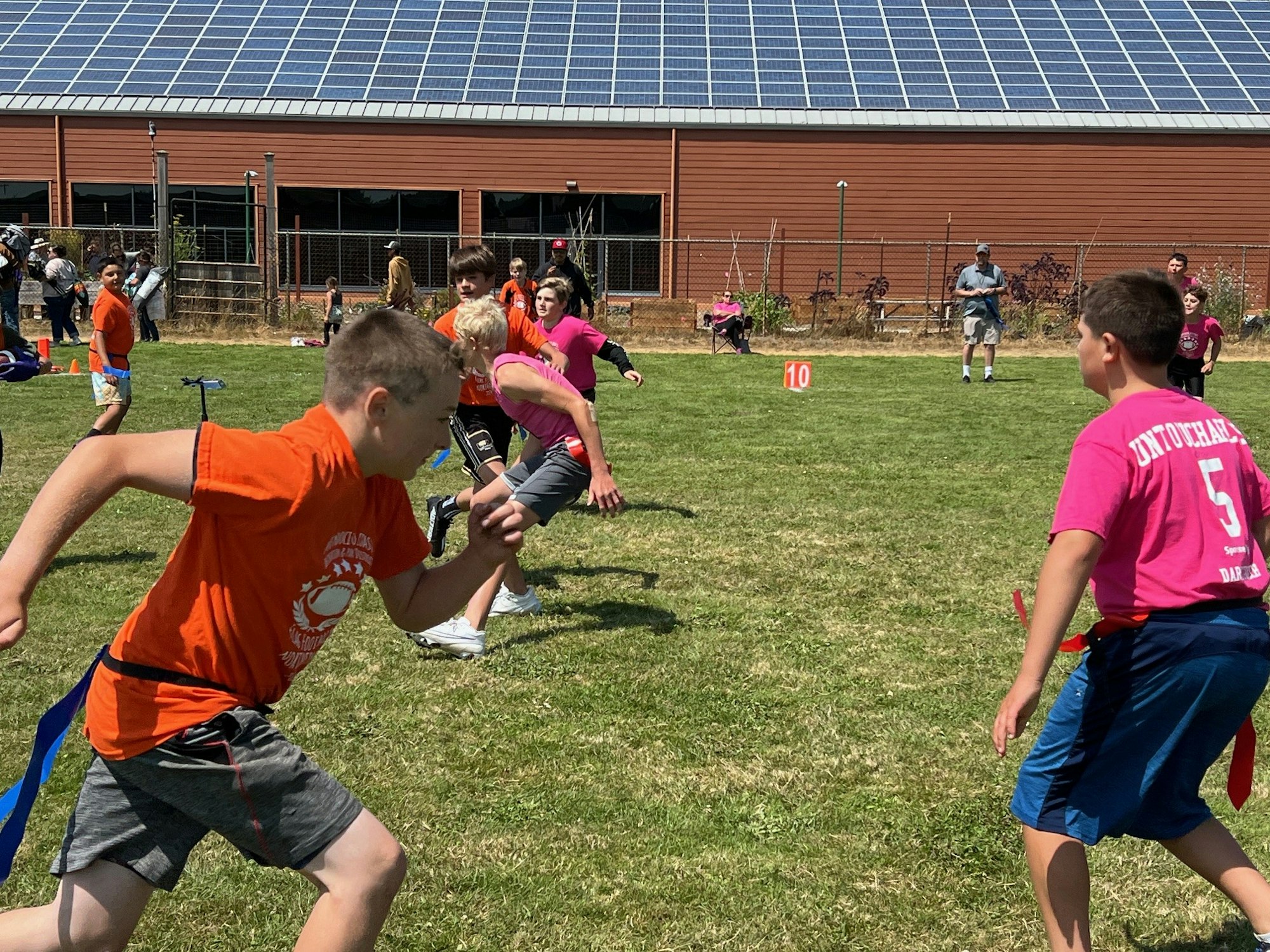 Children playing a flag football game on a grassy field with a building featuring solar panels in the background.