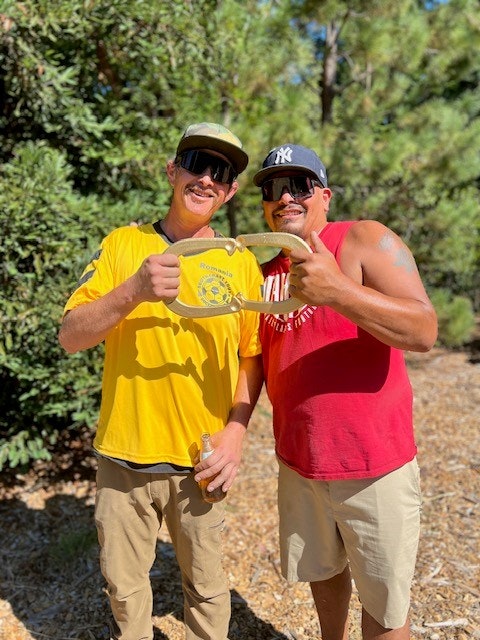 Two smiling men holding a belt, wearing caps and sunglasses, standing outdoors with trees in the background.