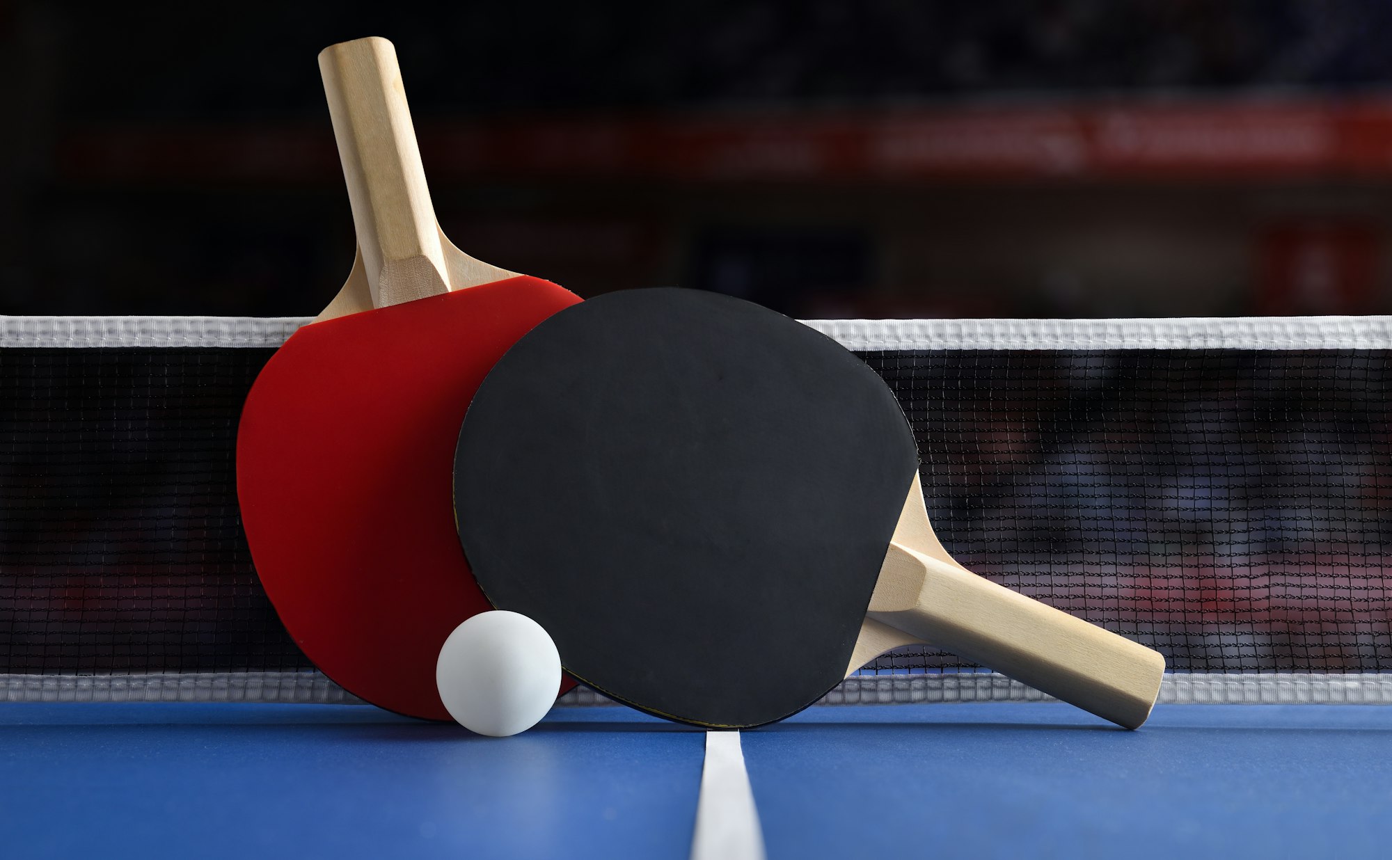 Two ping pong paddles, one red and one black, with a white ball on a blue table tennis surface, near a net.