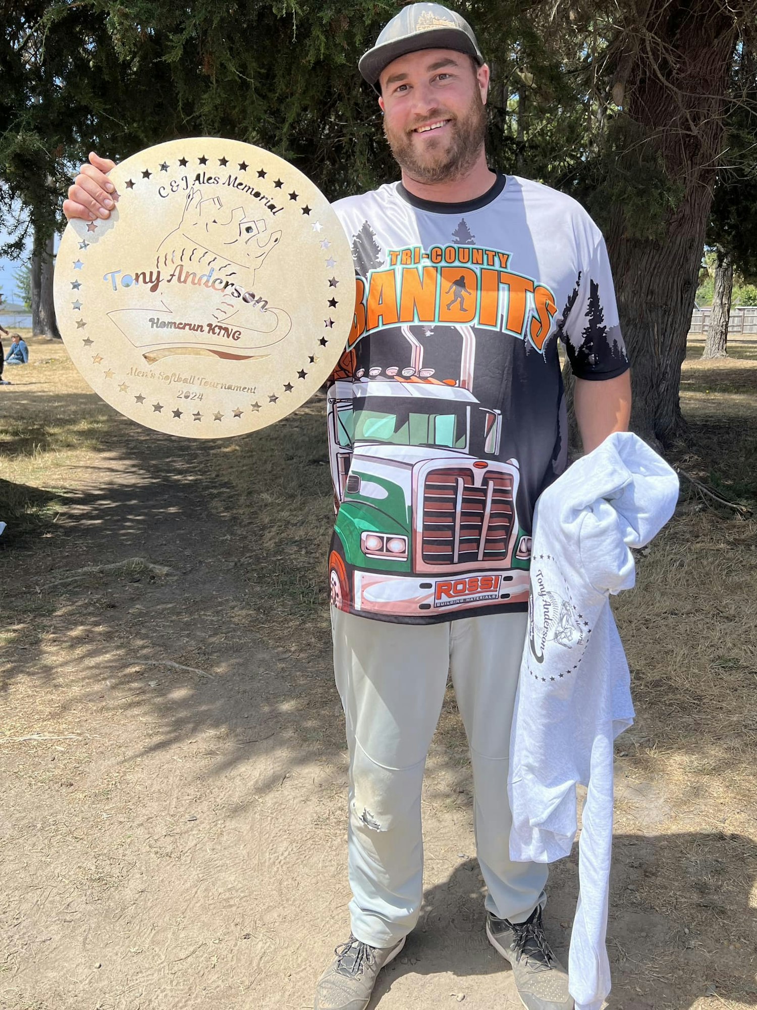 A man holds a round memorial plaque and wears a baseball shirt with a truck design.