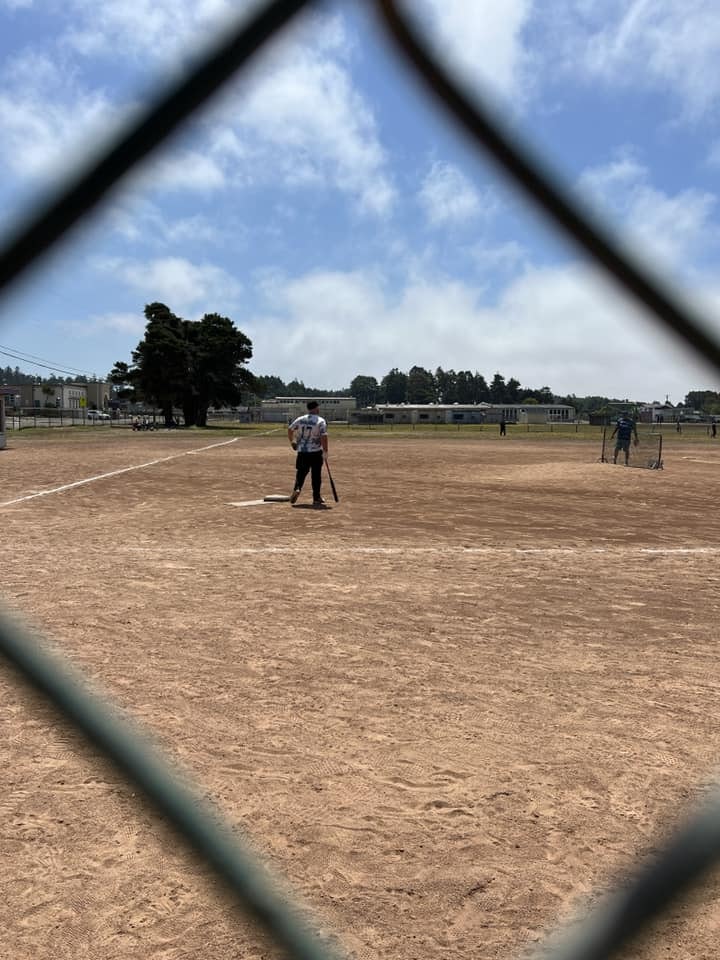 A baseball field viewed through a fence, with a person standing on the diamond and others in the background.