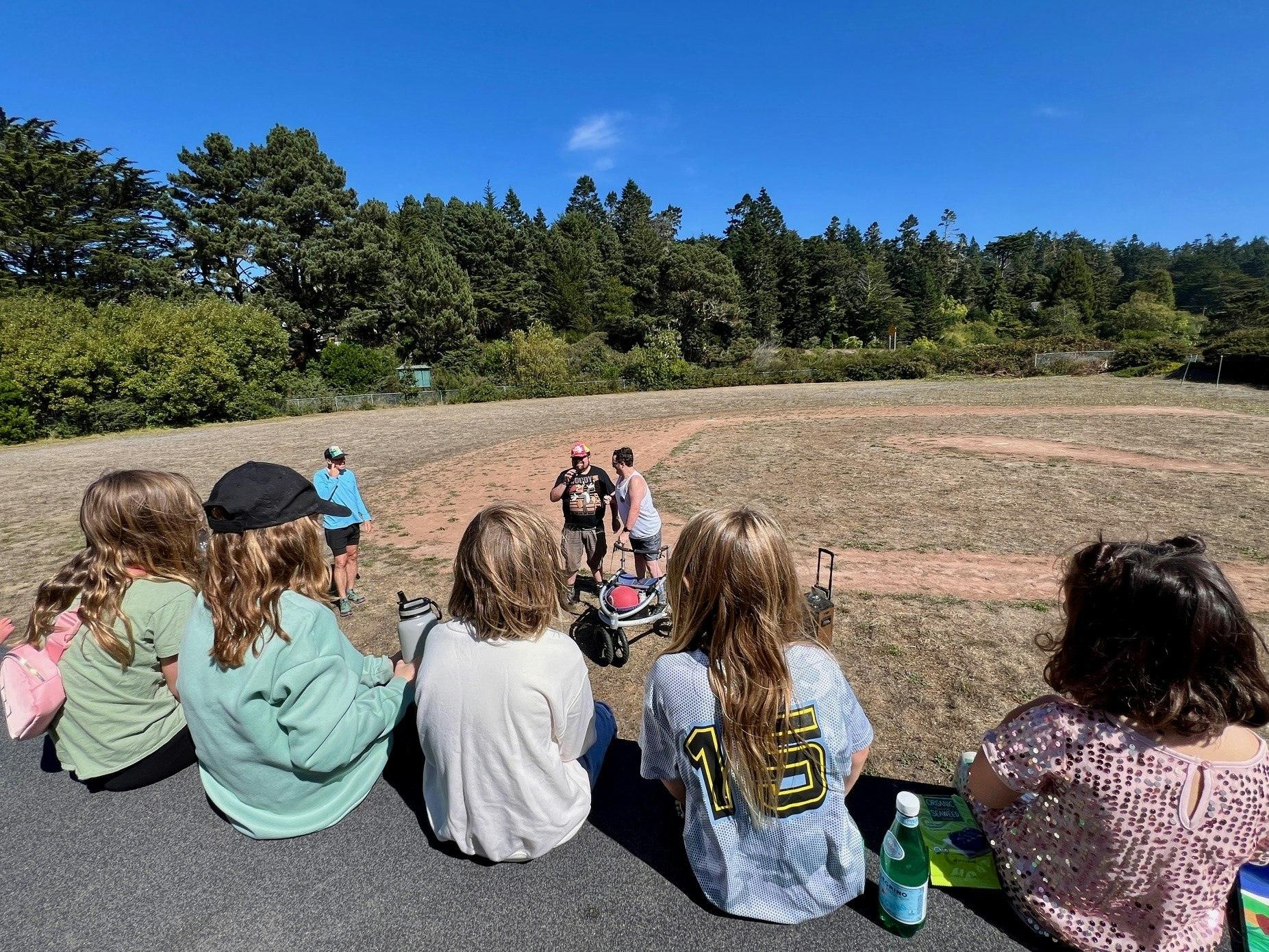 People sitting on a ledge watching others in a sunny, tree-surrounded field.