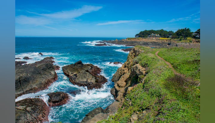 Coastal scene with rocky cliffs, ocean waves, grassy path, and distant house under a clear blue sky.