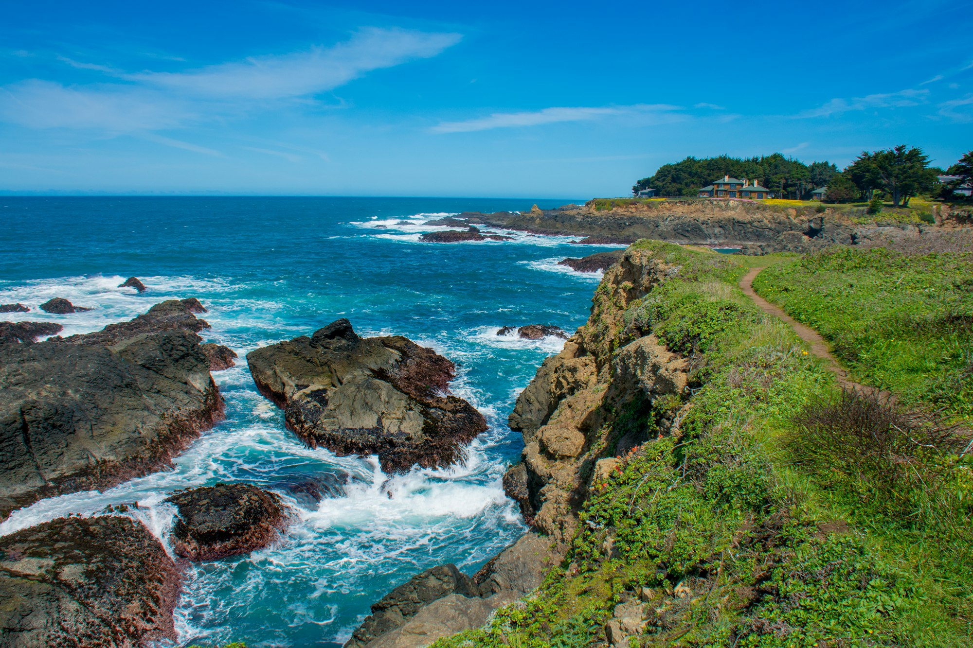 Coastal scene with rocky cliffs, ocean waves, grassy path, and distant house under a clear blue sky.