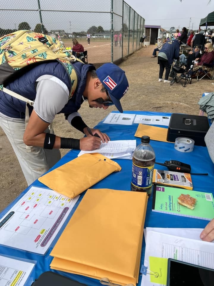 A person is signing a document at a table with papers and personal items, at an outdoor event.
