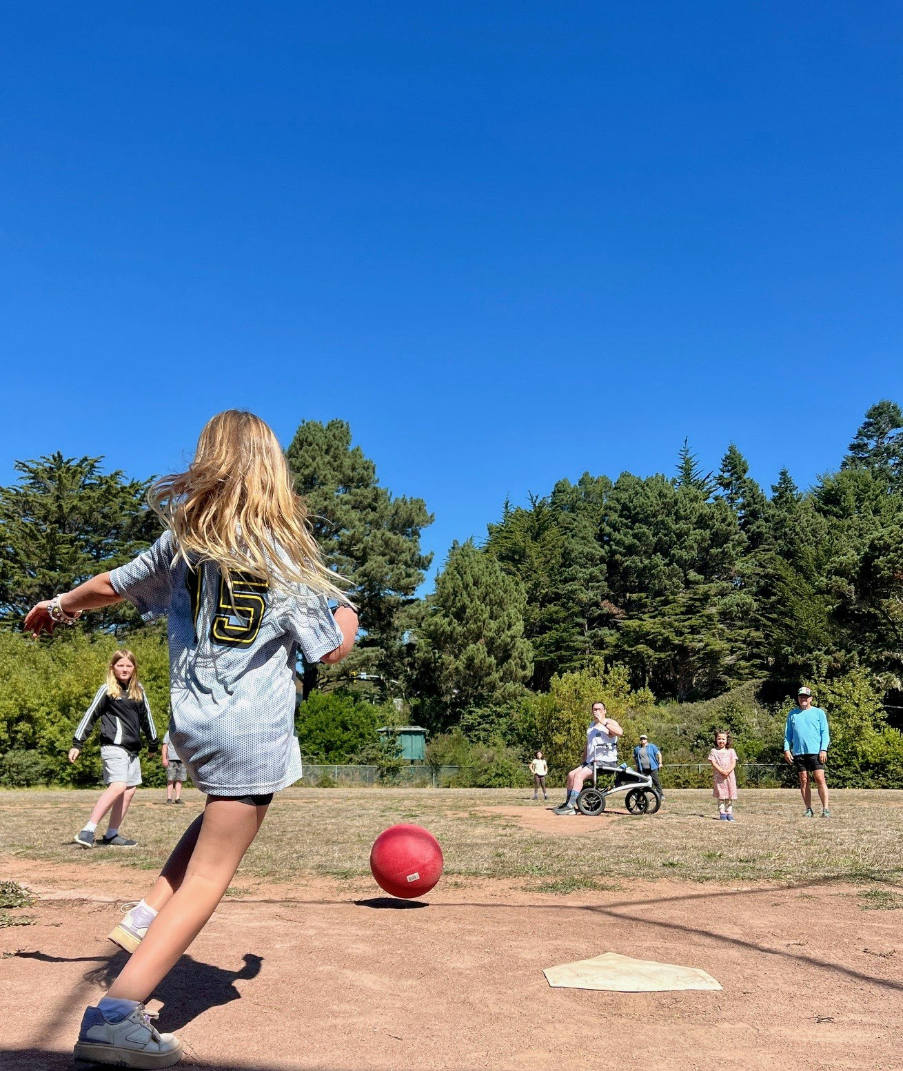 A group of people outdoors with a girl playing kickball in the foreground.
