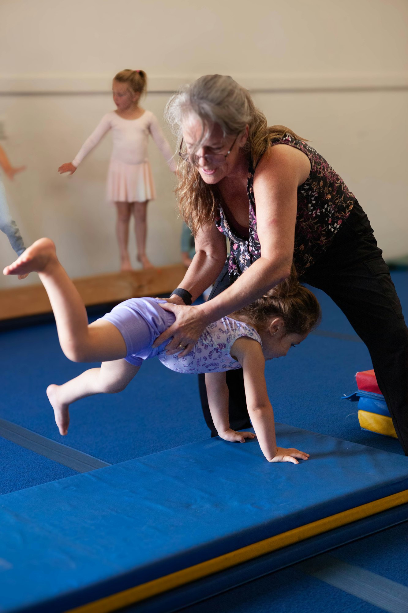 An adult assisting a child with gymnastics in a gym while another child watches in the background.